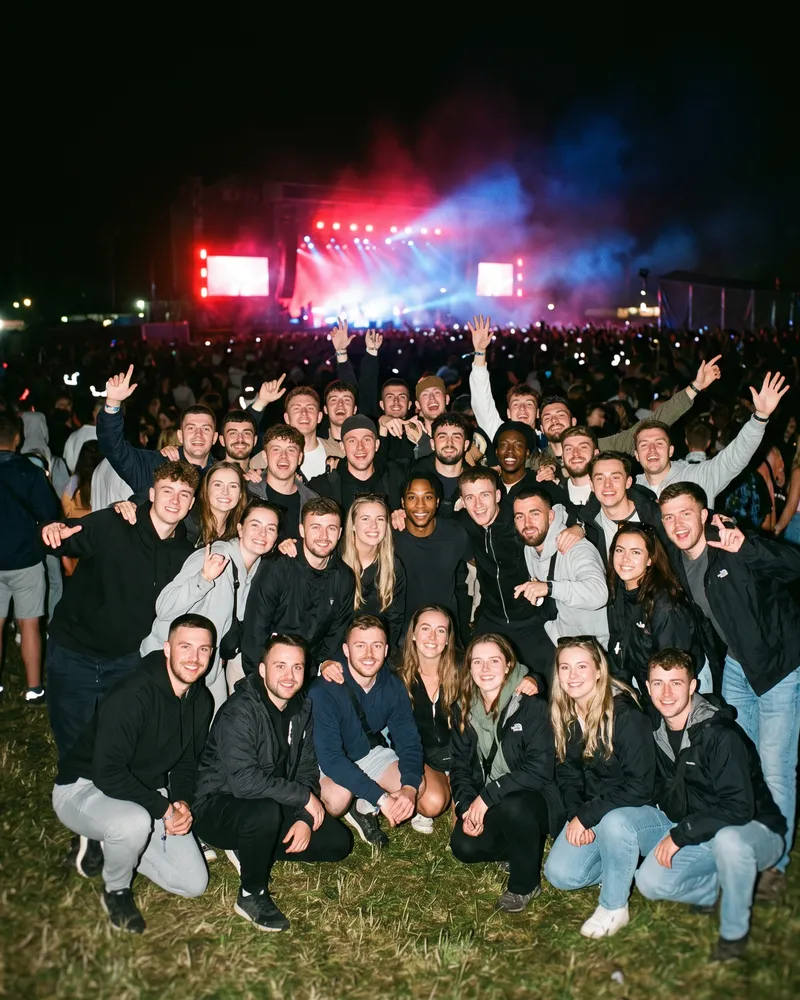 The same man standing small in the middle rows of a large group photo taken at a night-time outdoor concert.